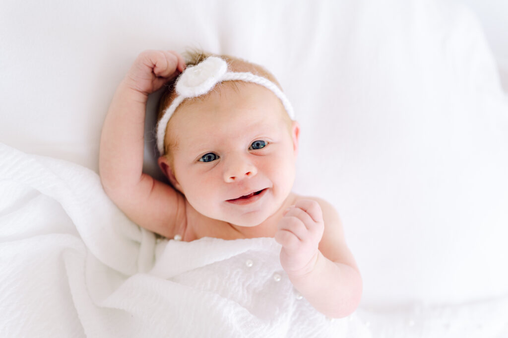 studio photoshoot of newborn girl wearing a white headband and a white blanket with pearls leaying over her